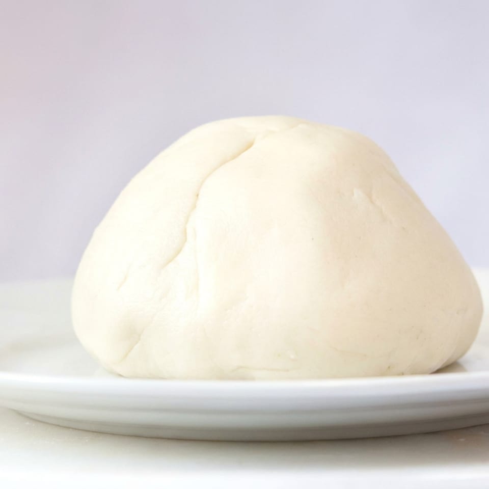 A smooth mound of pale dough sits on a white plate against a softly lit background.